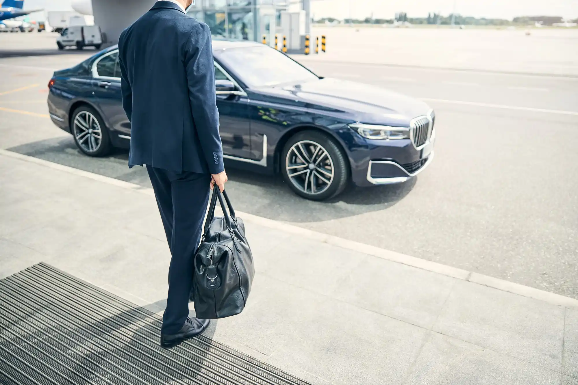 sharply dressed business traveler stepping out of a black executive sedan at a Boeing Field FBO entrance, rolling carry-on and leather briefcase visible, glass terminal reflecti...