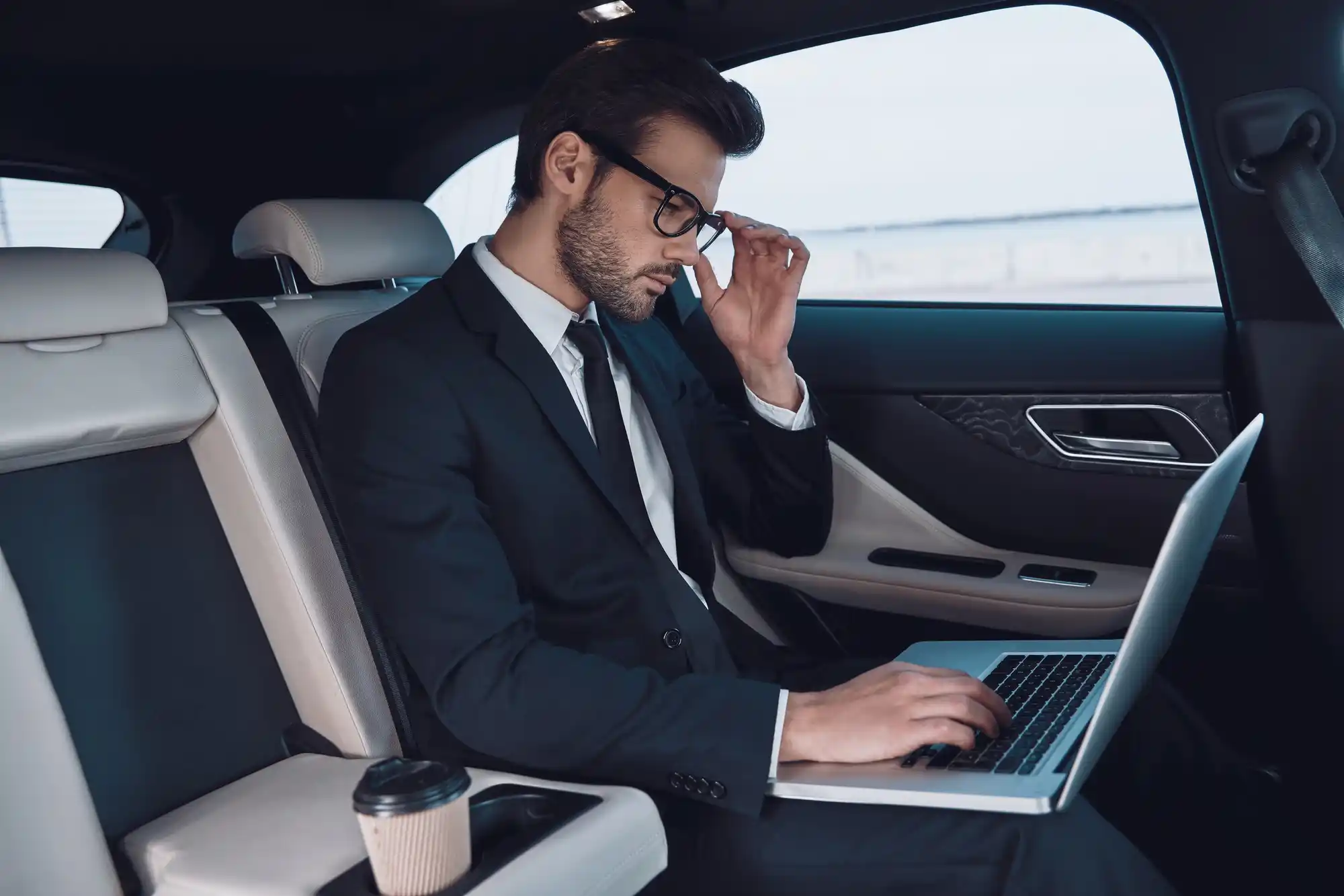 business traveler in navy suit working on laptop in rear seat of executive town car, Seattle freeway visible through rain-speckled window, soft ambient cabin lighting