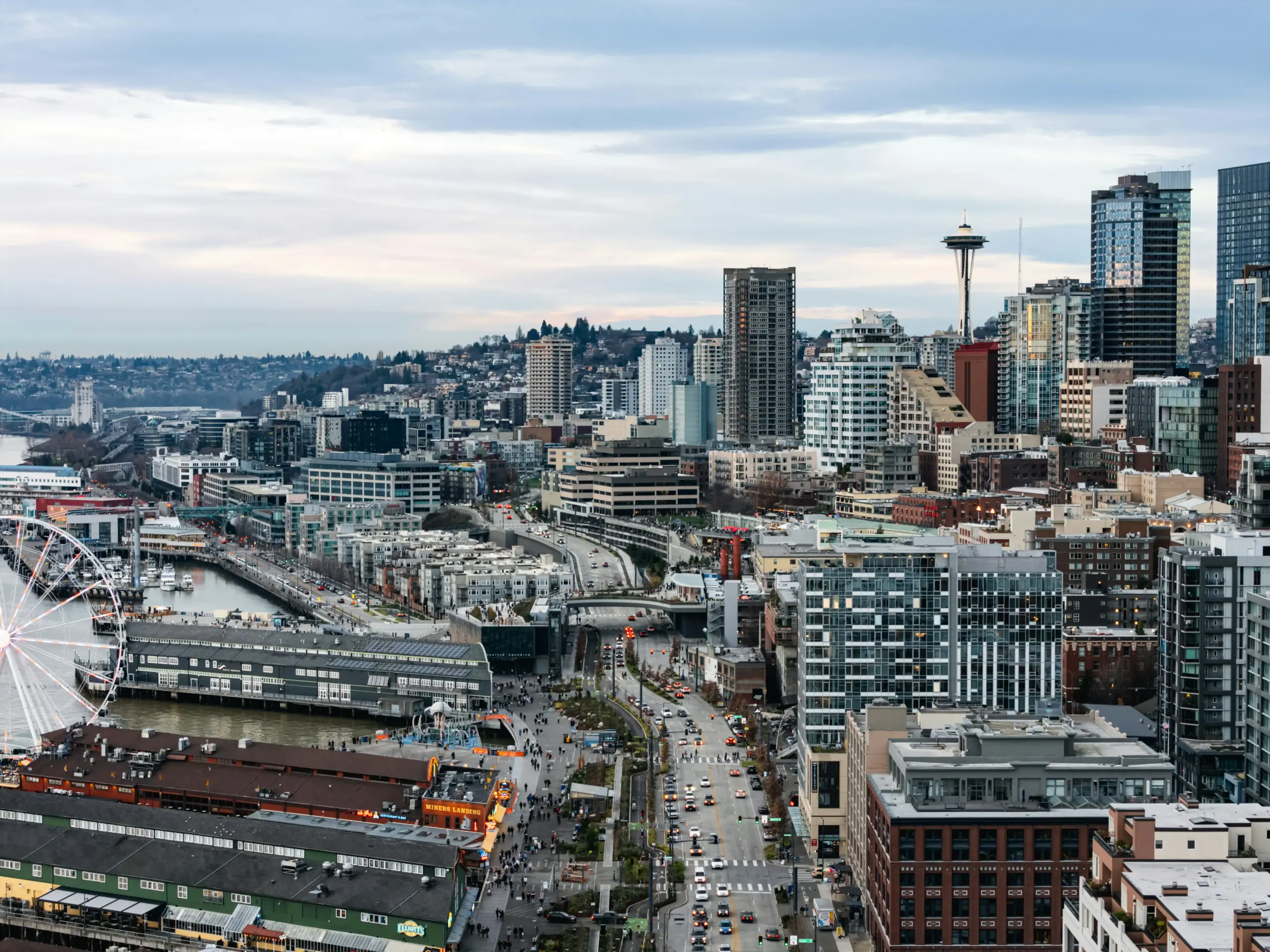 corporate travel coordinator at modern office desk in Seattle, dual monitors showing calendar and transportation bookings, coffee cup, city skyline visible through large windows