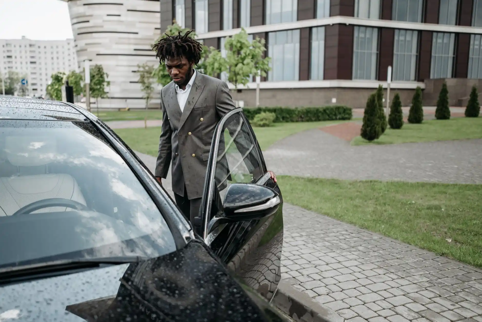 glossy black luxury sedan parked curbside outside a modern downtown Seattle hotel at dusk, chauffeur in dark suit opening the rear door, city lights reflecting on wet pavement
