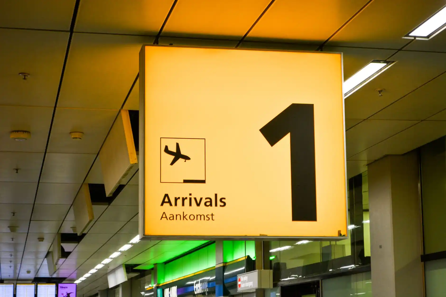 professional chauffeur in dark suit holding a digital name sign inside Sea-Tac arrivals area, polished black sedan visible through glass, warm terminal lighting and business tra...
